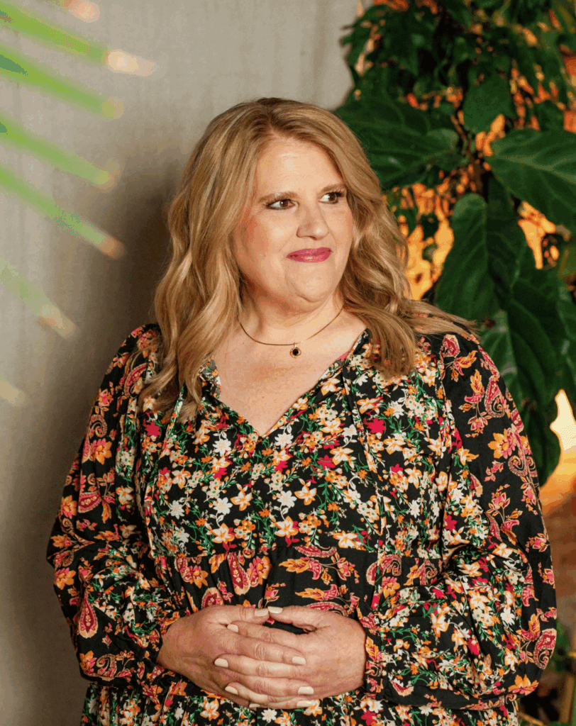 Woman smiling in floral dress with lush green plants in background.