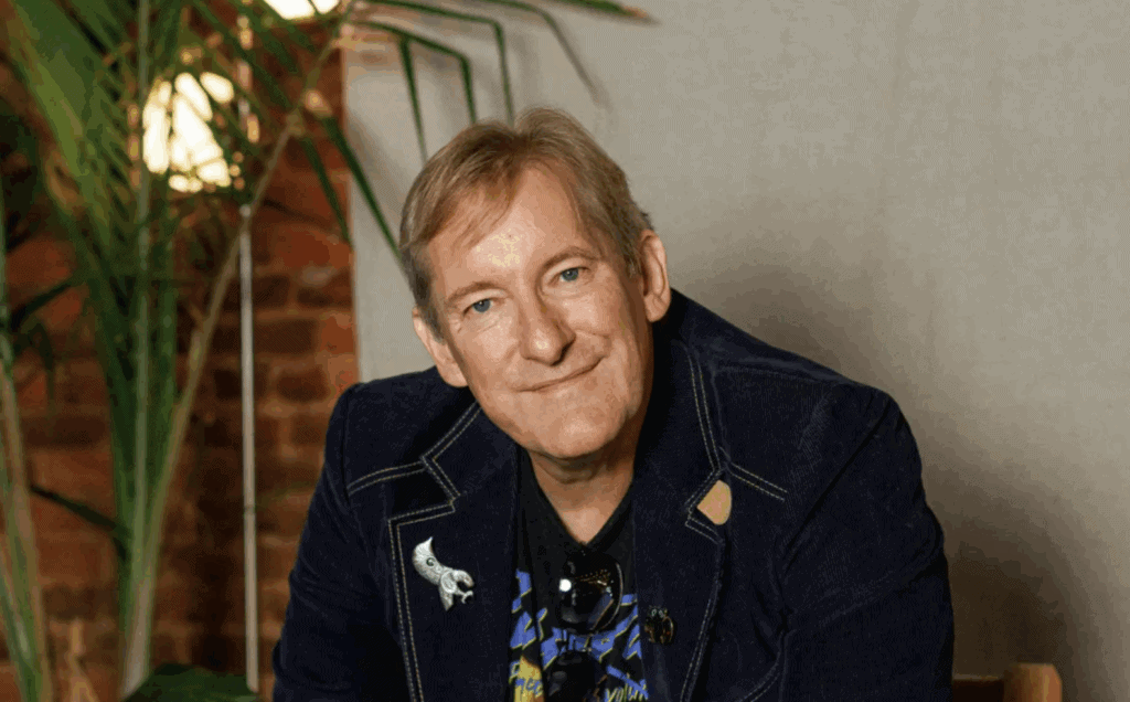 Man smiling, sitting indoors with greenery and brick wall background.