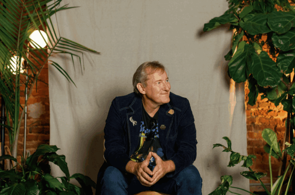 Elderly man sitting among lush green plants in cozy indoor setting.