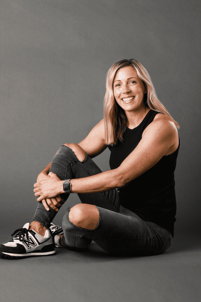 Relaxed woman sitting on the floor with a smile, wearing casual black outfit and sneakers.