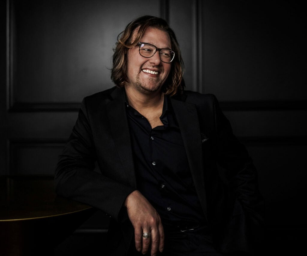 man with shoulder length hair smiles in all black on black wall