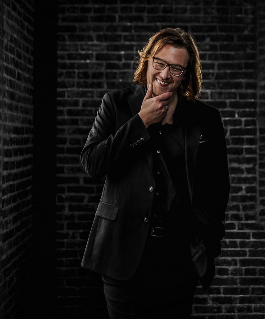 man with shoulder length hair smiles in all black on black wall