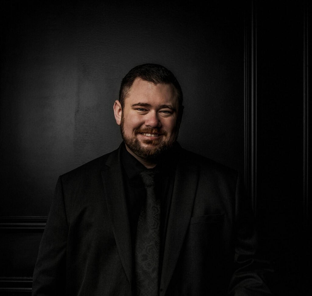 Man in black suit smiling, standing against a dark background, representing local business and commu.