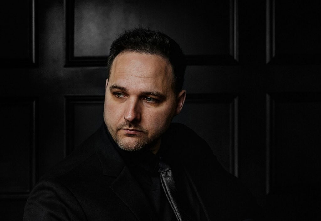 Man in black suit with serious expression, dark background, professional portrait.