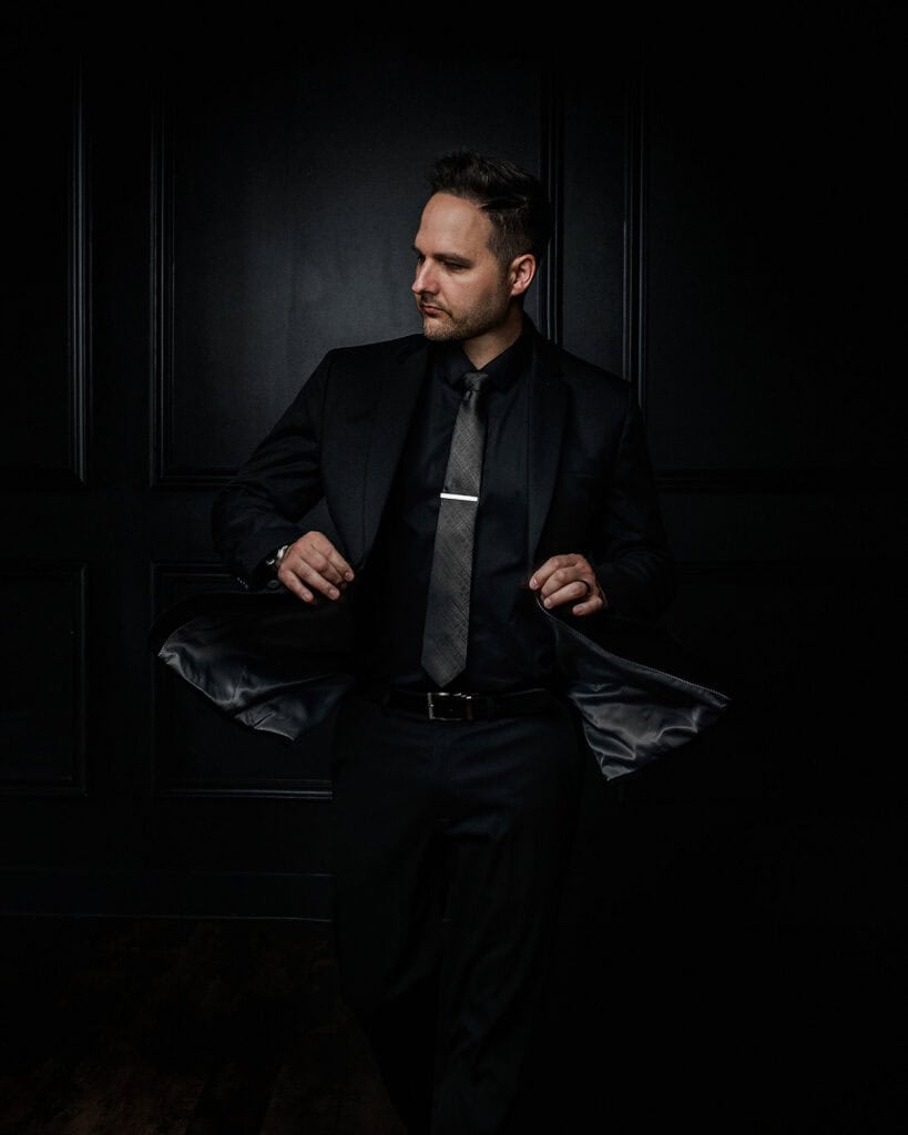 Elegant man in black suit with a tie, standing confidently in a dark setting.