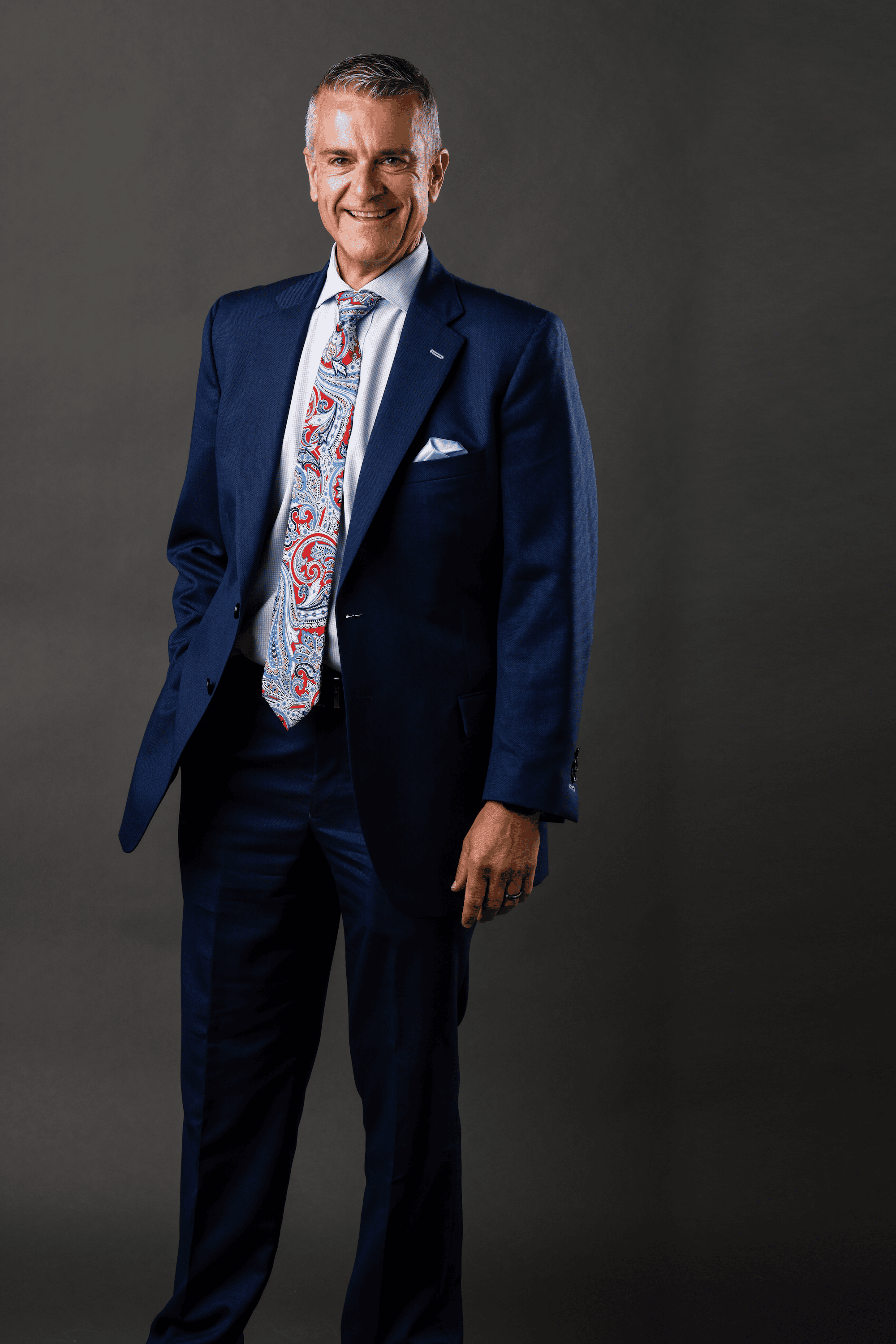 Businessman in a navy suit with a colorful tie, standing confidently against a gray background.